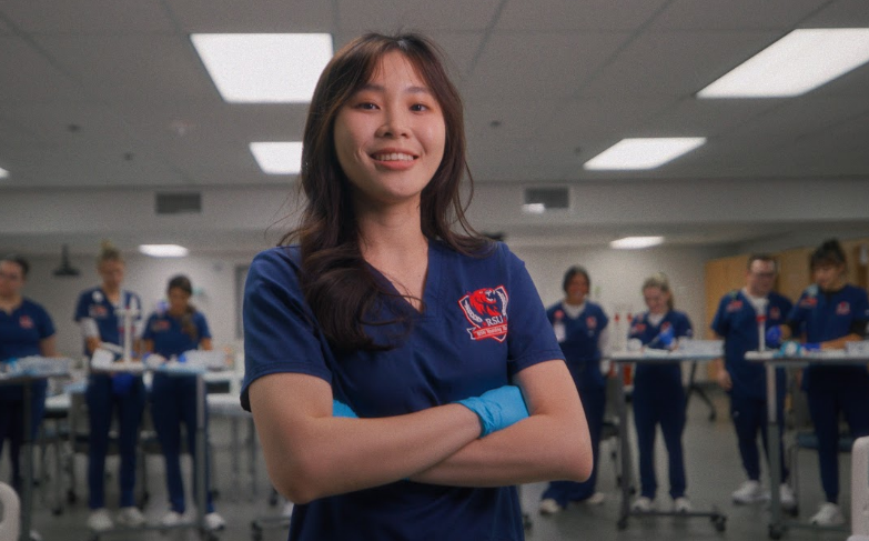 A female nursing student in blue scrubs standing in front of other students at tables.